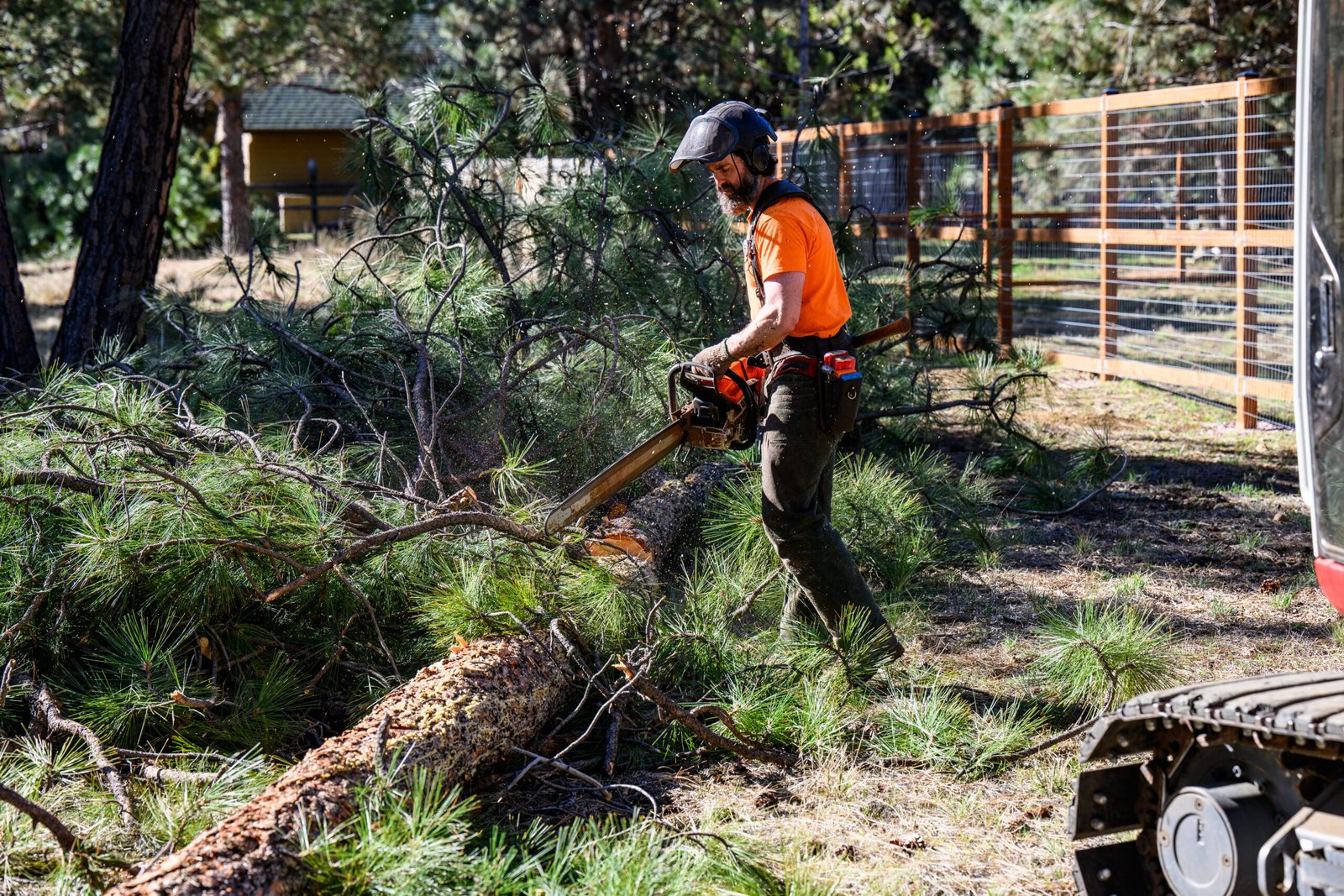 A forestry worker wearing safety gear uses a chainsaw to cut a fallen pine tree into sections on a wooded Central Oregon property. Pine branches and slash are scattered on the ground near a fence, with forestry equipment visible in the foreground during fuels reduction and debris cleanup work.
